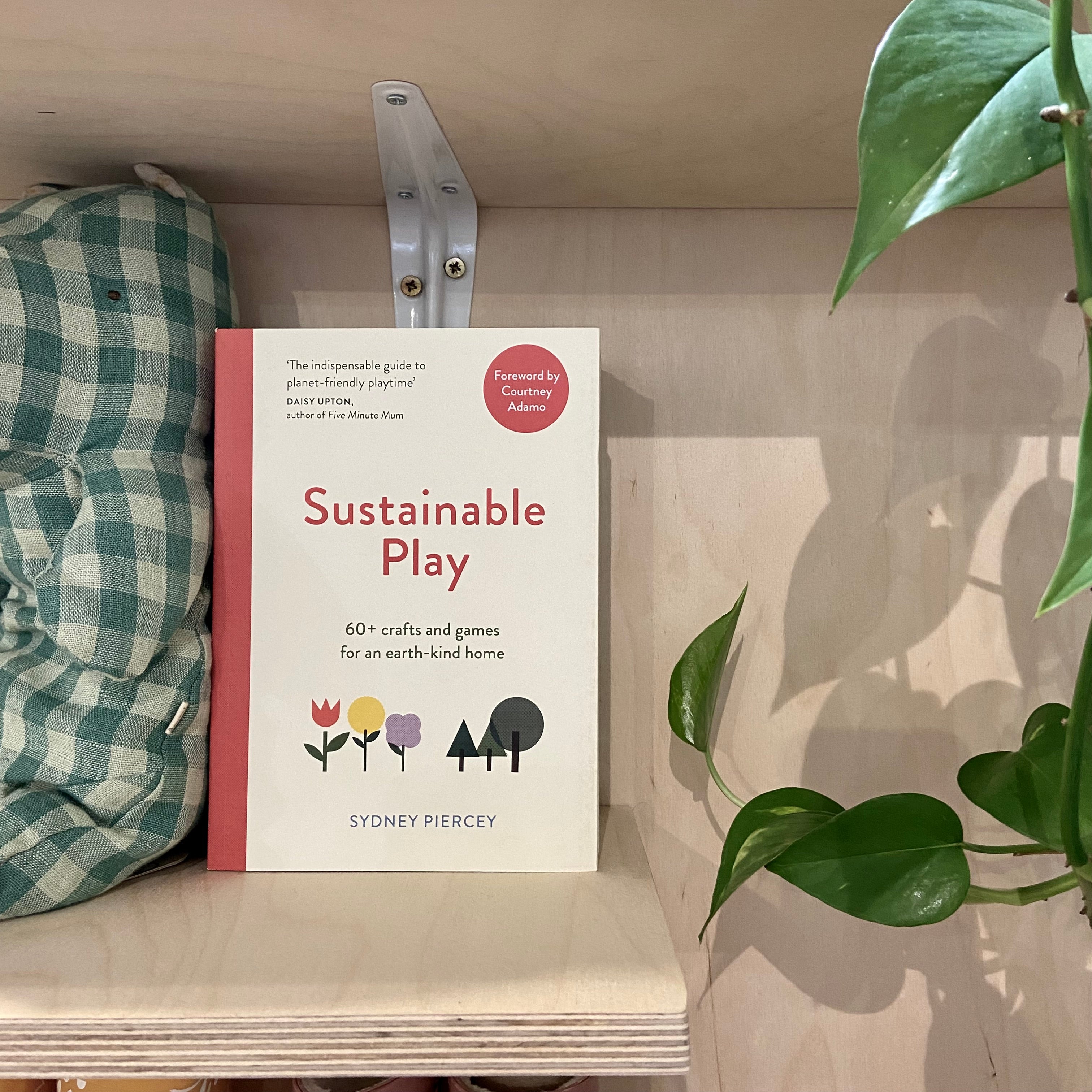 A book titled 'Sustainable Play' is placed on a shelf alongside a green checked hippo and green foliage on the other side of the image.